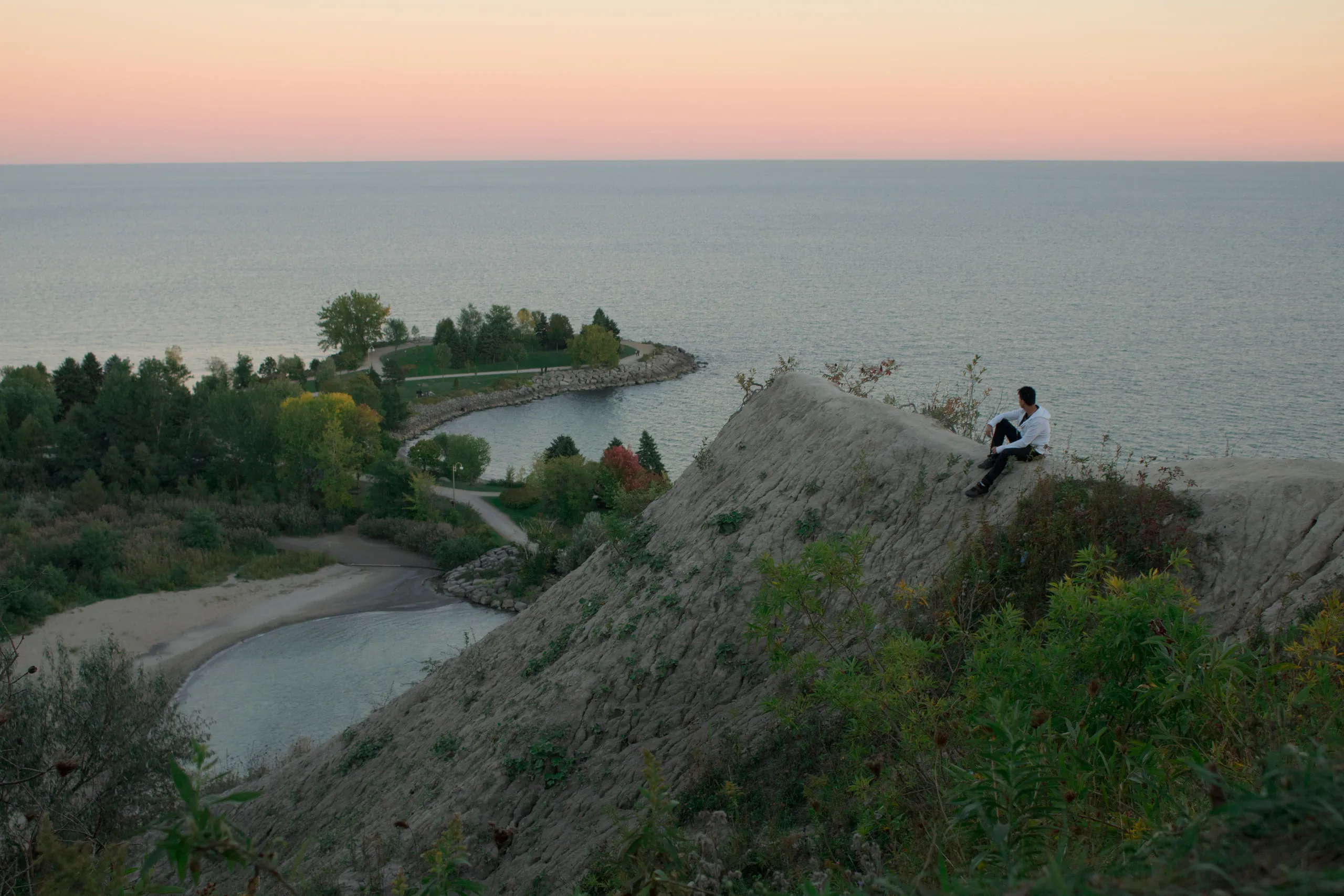A person sits on a bluff overlooking the lake beneath a pastel sunset.
