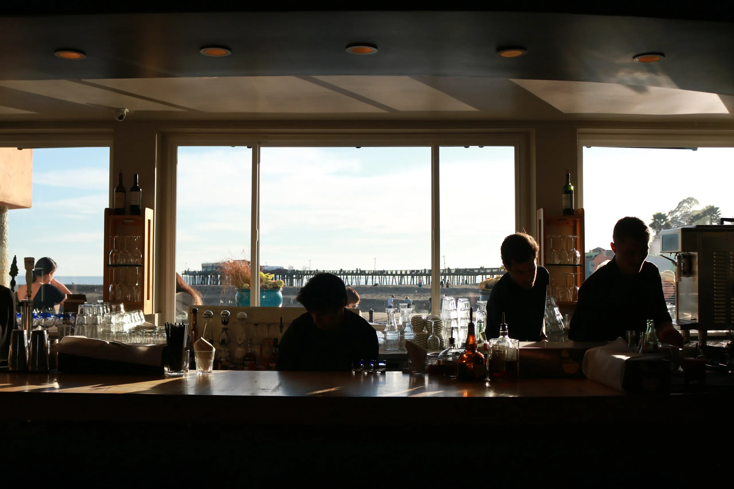 Restaurant staff in silhouette against windows overlooking the coast and a pier.
