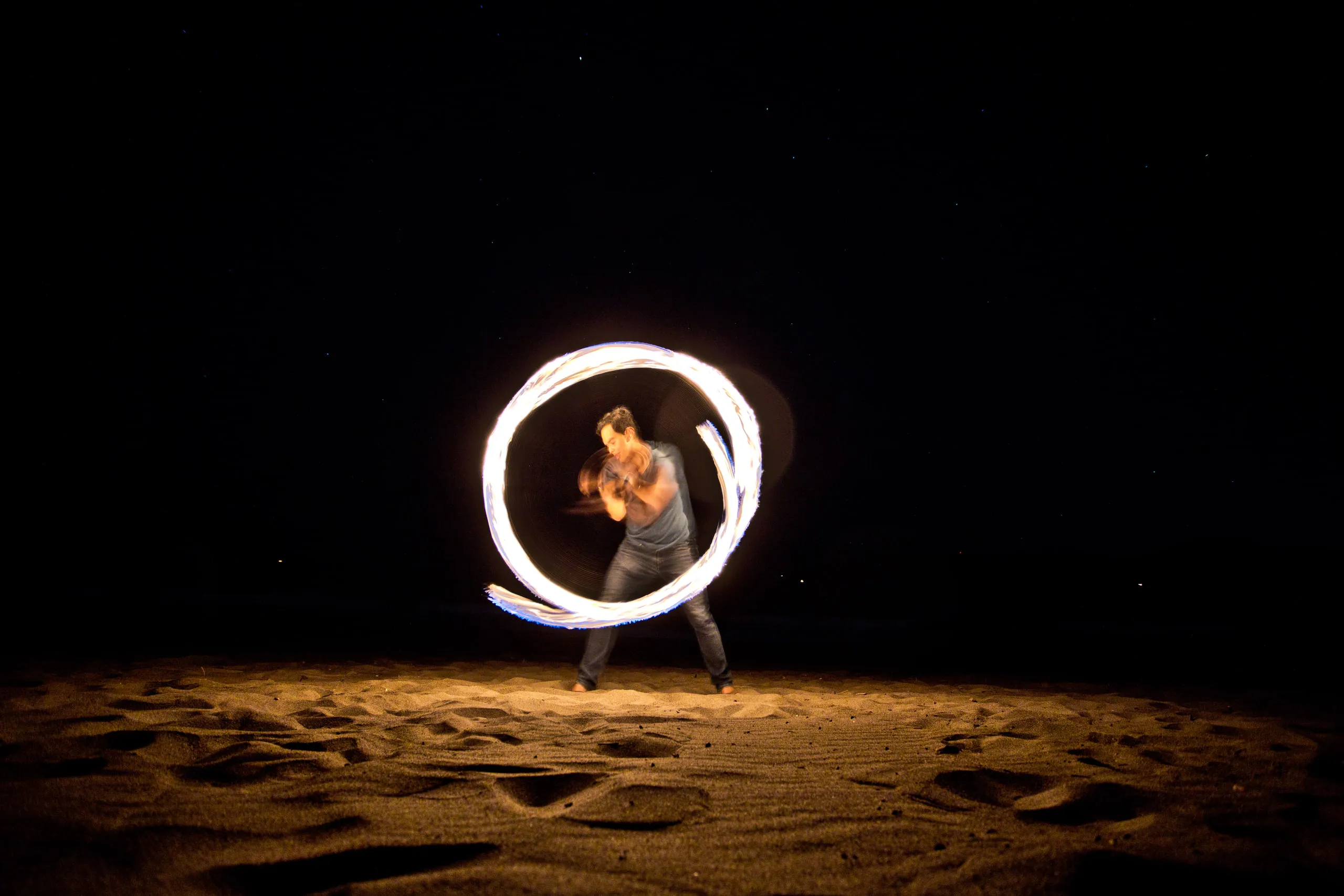 A fire performer spins a bright circle of light on a dark beach.